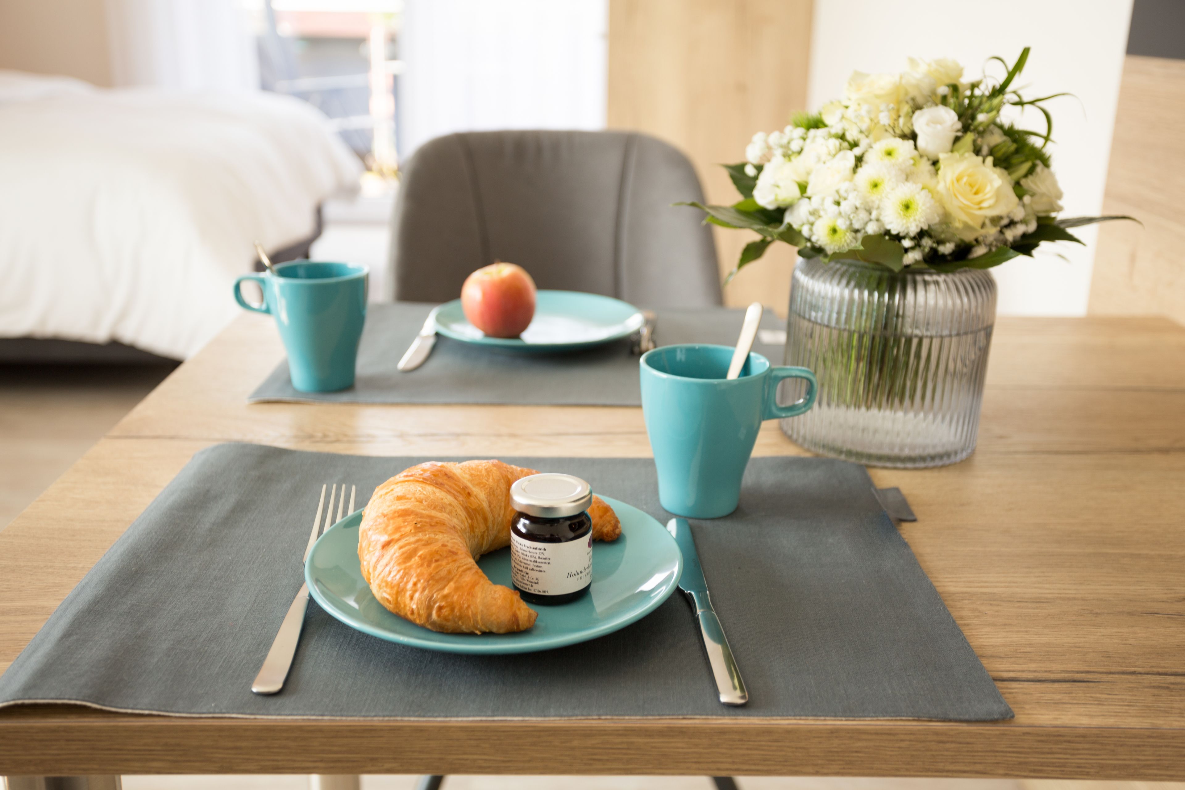 Morgenmad med croissant, æble og kaffe på et bord, en vase med blomster, og en seng i baggrunden.