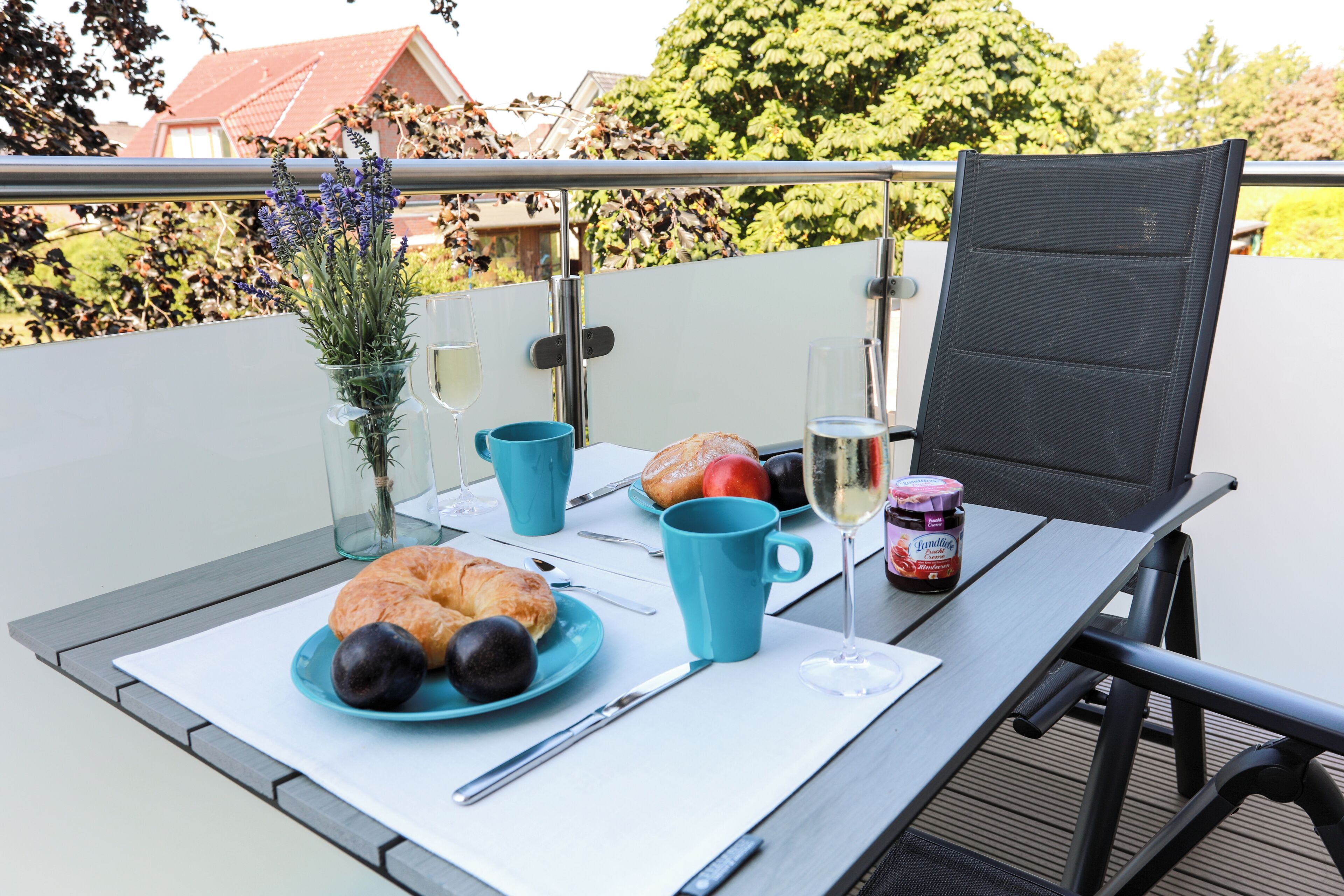 Een tafel met croissants, fruit, een glas wijn en een pot jam op een balkon met uitzicht op bomen en huizen.