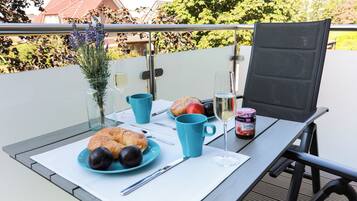 Een tafel met croissants, fruit, een glas wijn en een pot jam op een balkon met uitzicht op bomen en huizen.