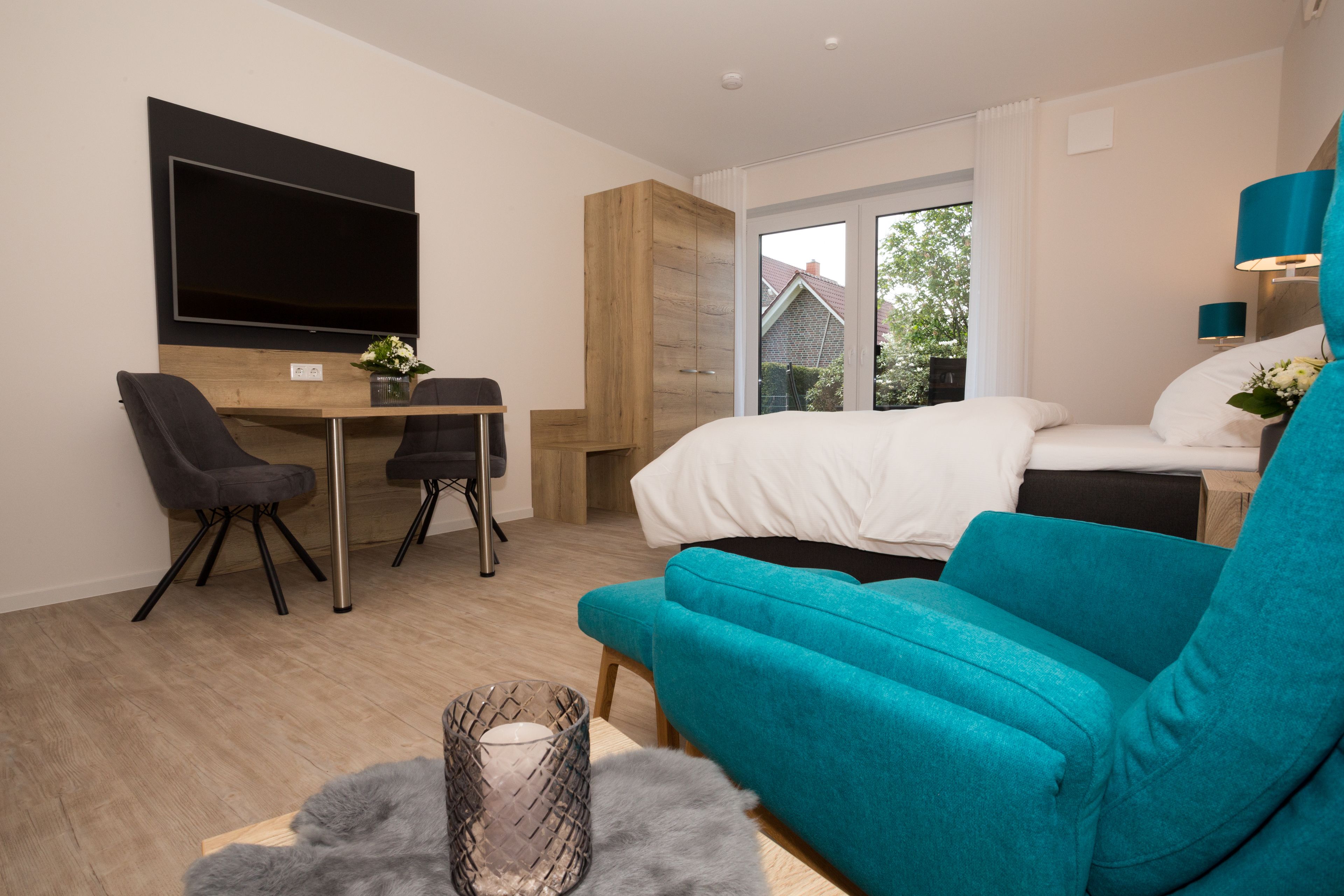 A modern hotel room with a large bed, a sofa, a desk with a chair, a flat-screen TV, and a large window offering a view of the outside.