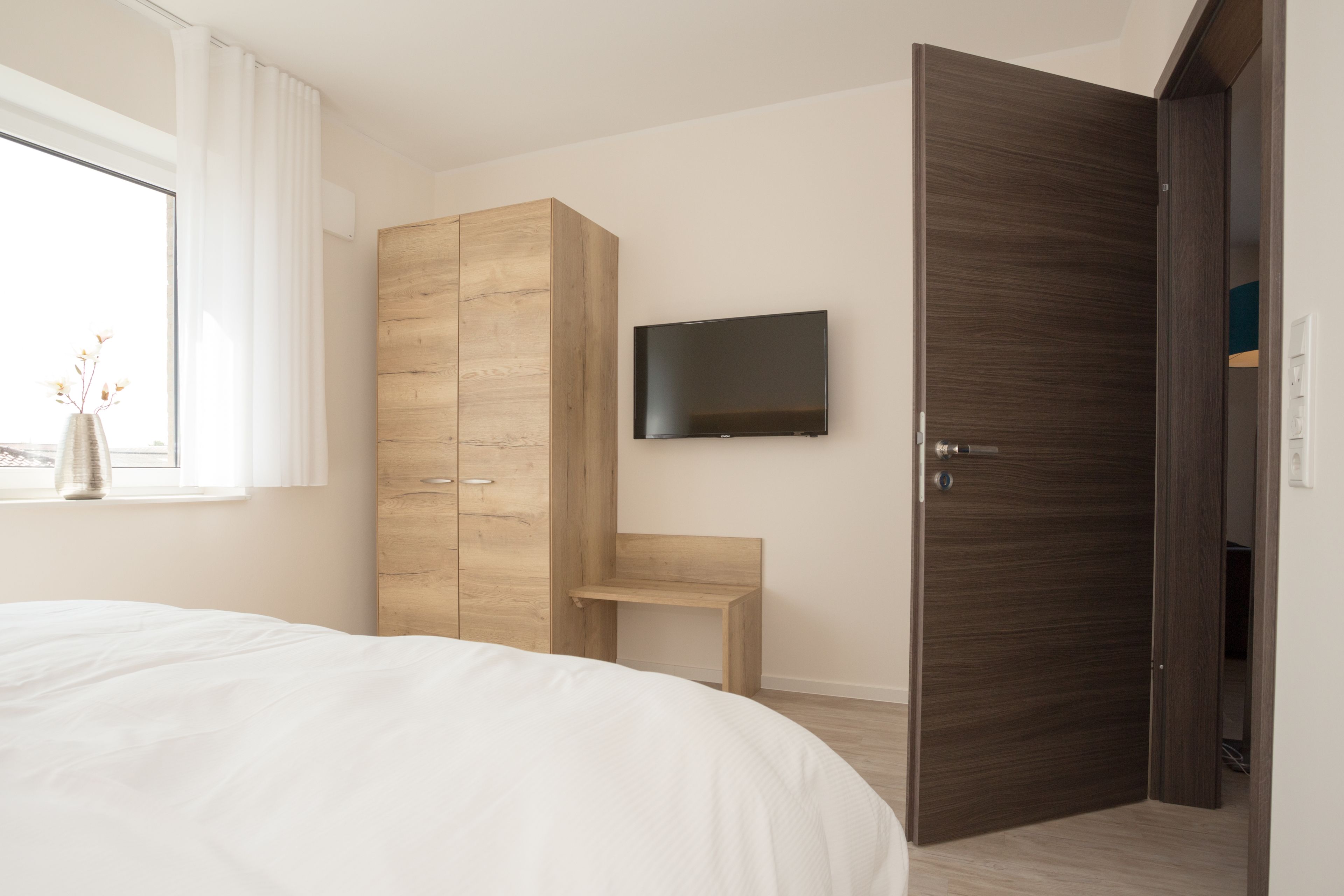 Un dormitorio con cama, un armario de madera, televisor montado en la pared y una puerta que da a otra habitación.