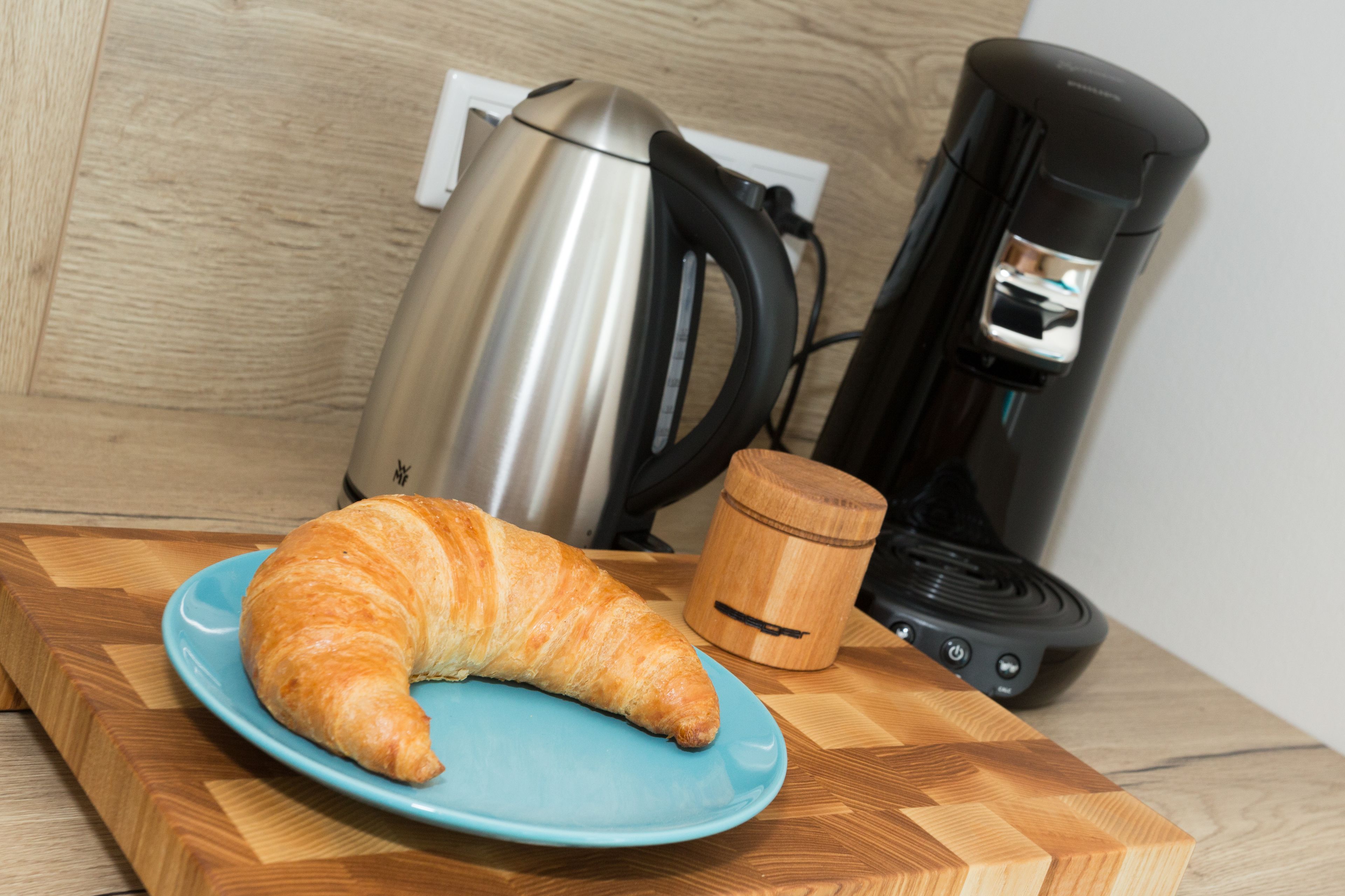 A croissant on a blue plate on a wooden cutting board, a stainless steel kettle, and a black coffee maker.