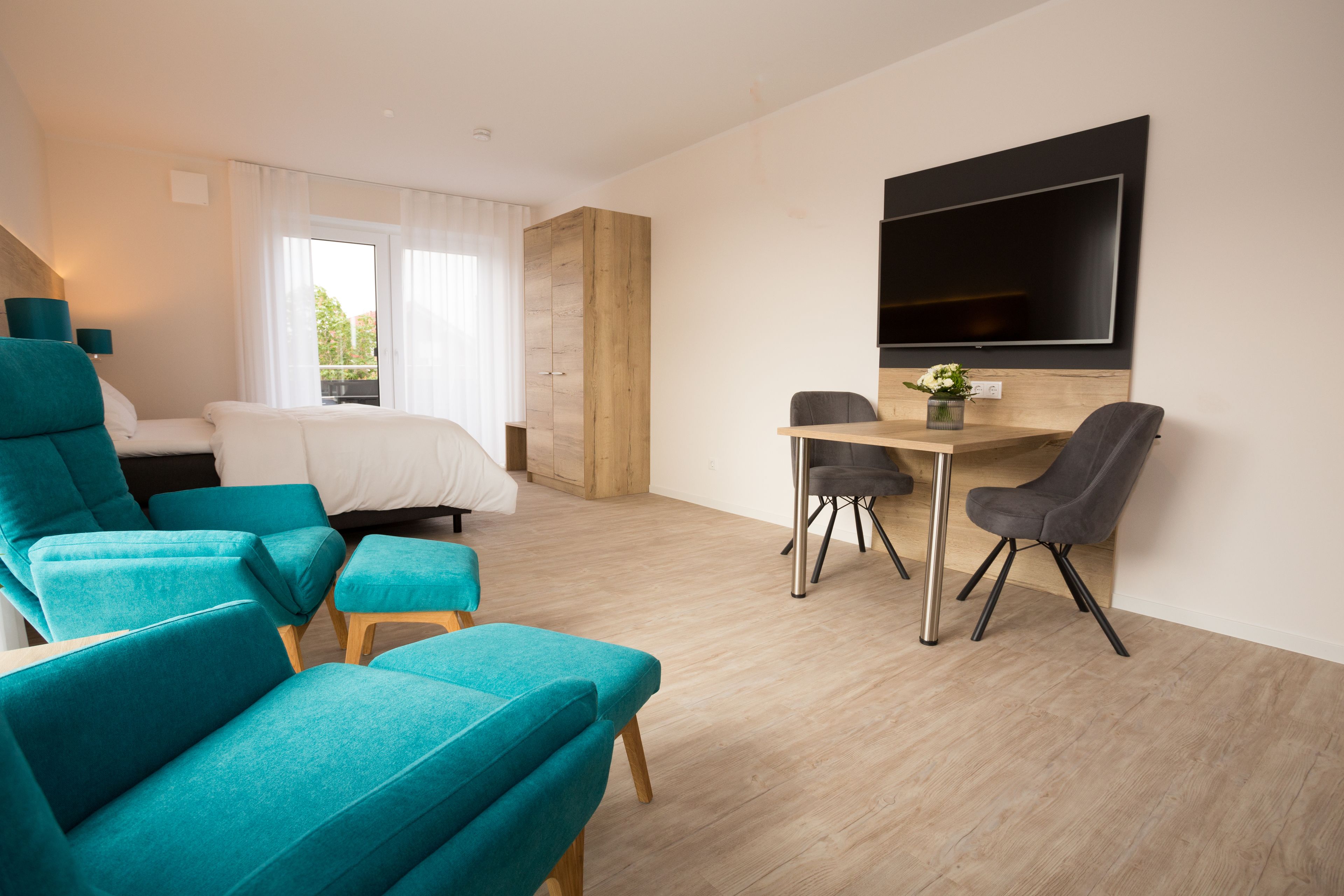 A modern hotel room with a bed, teal armchairs, a dining table with chairs, a TV mounted on the wall, and a wooden wardrobe.