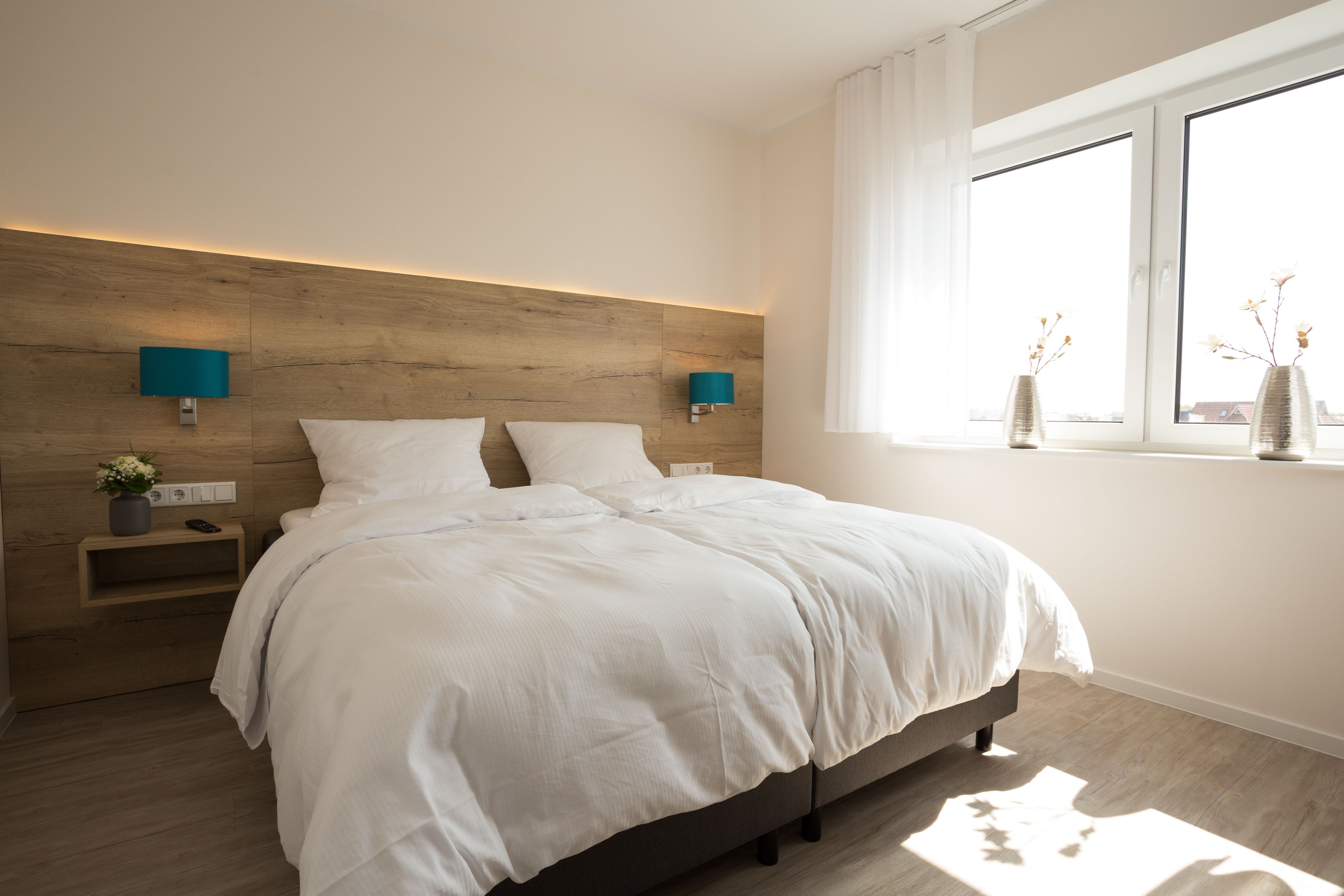 Un dormitorio moderno con cabecera de madera, una cama grande con ropa de cama blanca, dos mesitas de noche con lámparas y una ventana con vista a la ciudad.