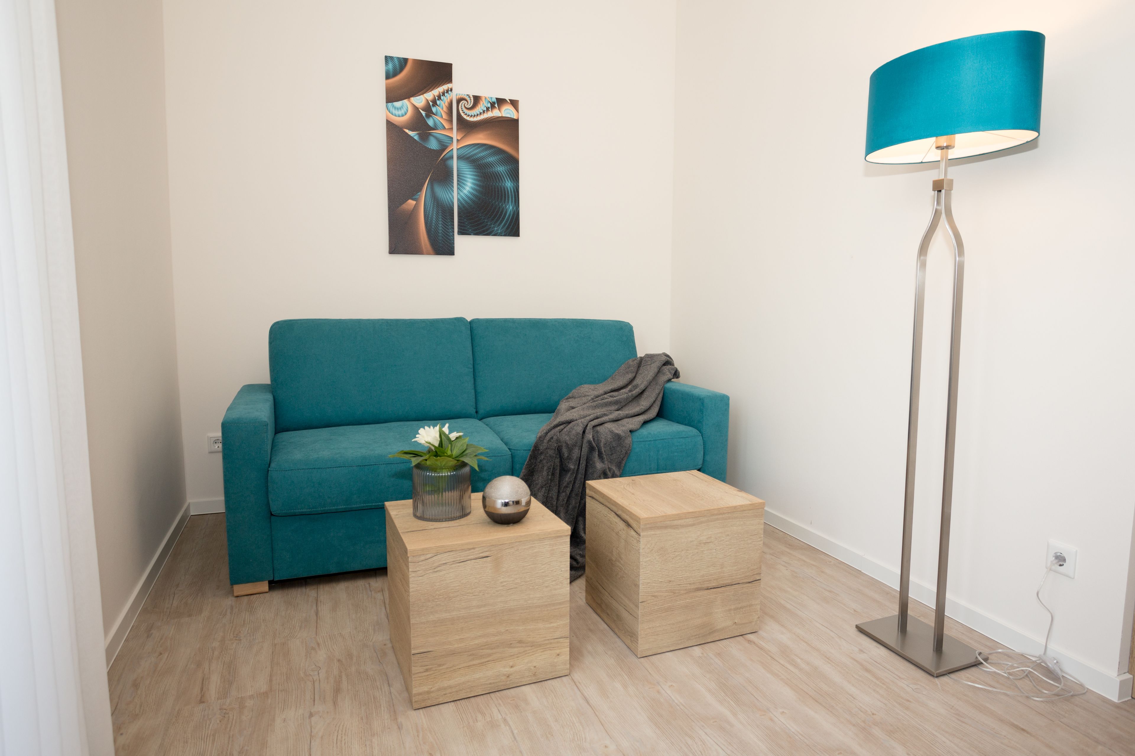A teal sofa with a blanket, two wooden side tables, a floor lamp, and abstract wall art.