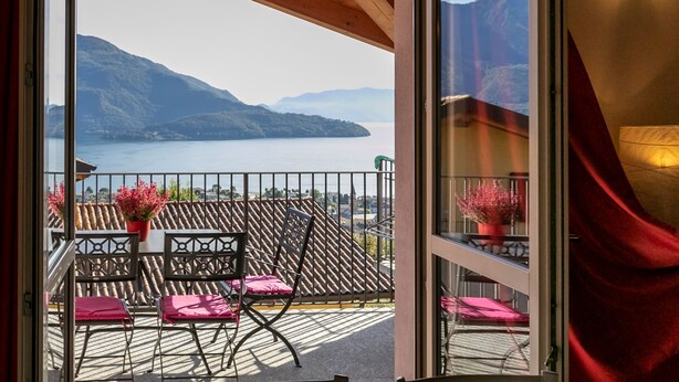 Un balcon ouvert donnant sur un lac et des montagnes. On y trouve deux chaises et une table avec un pot de fleurs.