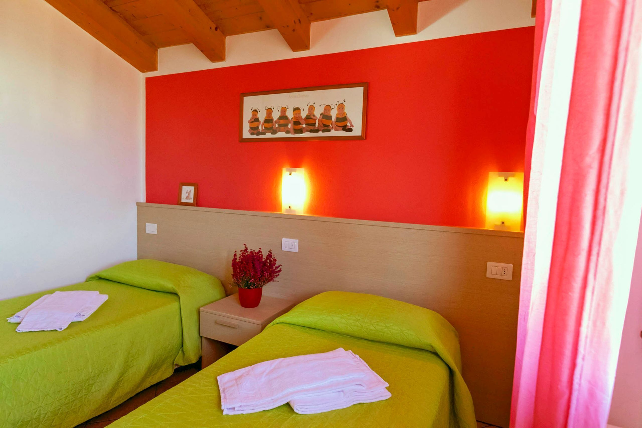 A hotel room with two single beds, each with a green bedspread and white towels. There is a small table with a red flower vase between the beds. The room has a red accent wall with a framed picture above the table.