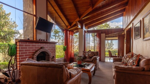 A spacious living area with a brick fireplace, leather sofas, and large windows offering a view of the outdoors. - Monteverde Panoramic House (Monteverde)