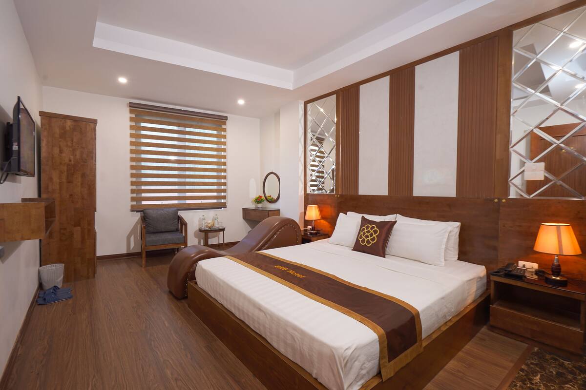 Une chambre d’hôtel avec un grand lit, une tête de lit en bois, un bureau avec une lampe, une chaise, une télévision et une fenêtre avec des stores.
