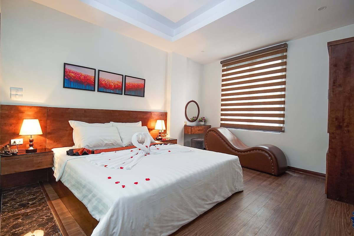 Une chambre d’hôtel comprenant un lit, deux lampes de chevet, une tête de lit en bois, un banc en bois, une grande fenêtre avec des stores et trois tableaux encadrés accrochés au mur.