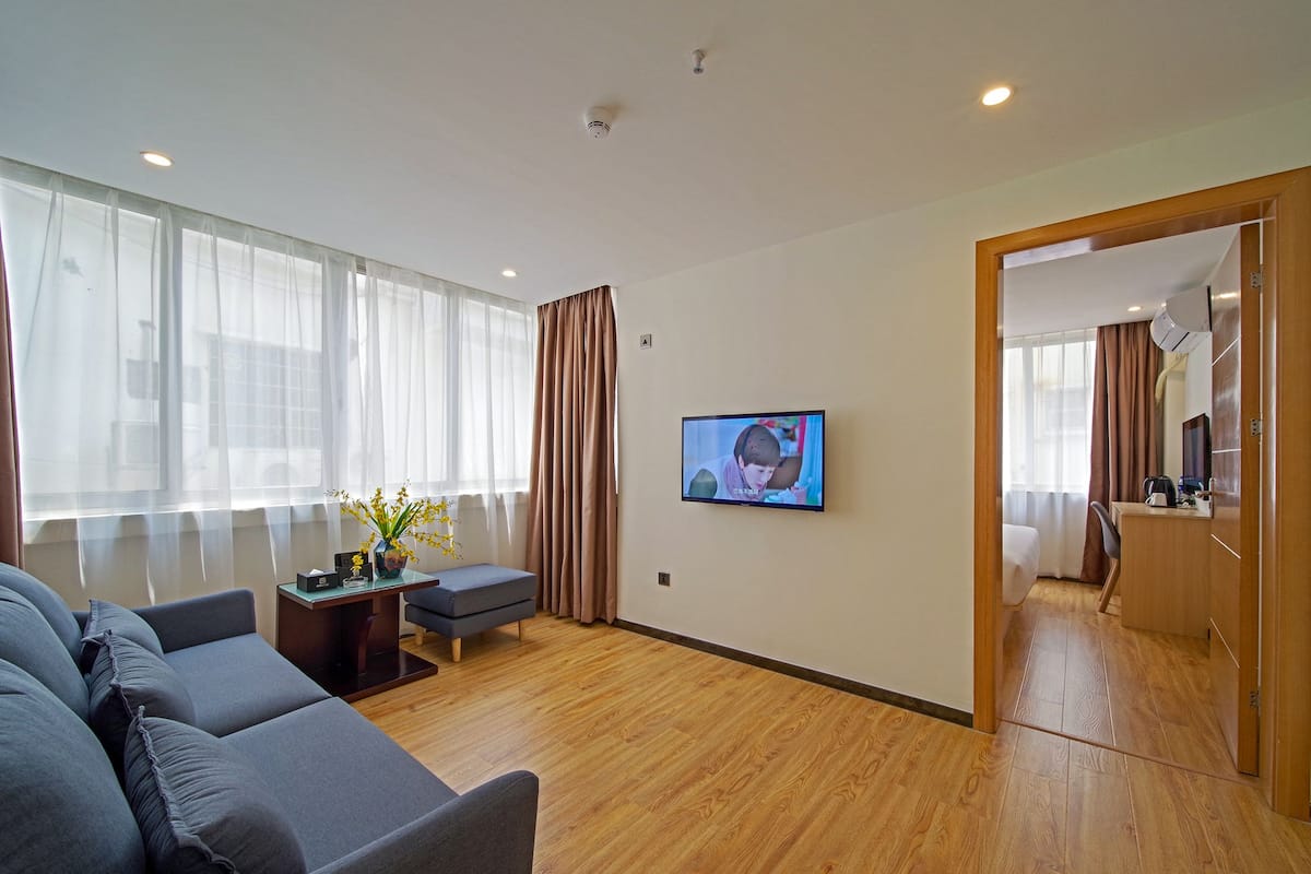 Una habitación de hotel moderna con un sofá grande, una mesita con un florero y una televisión montada en la pared, además de un suelo de madera.