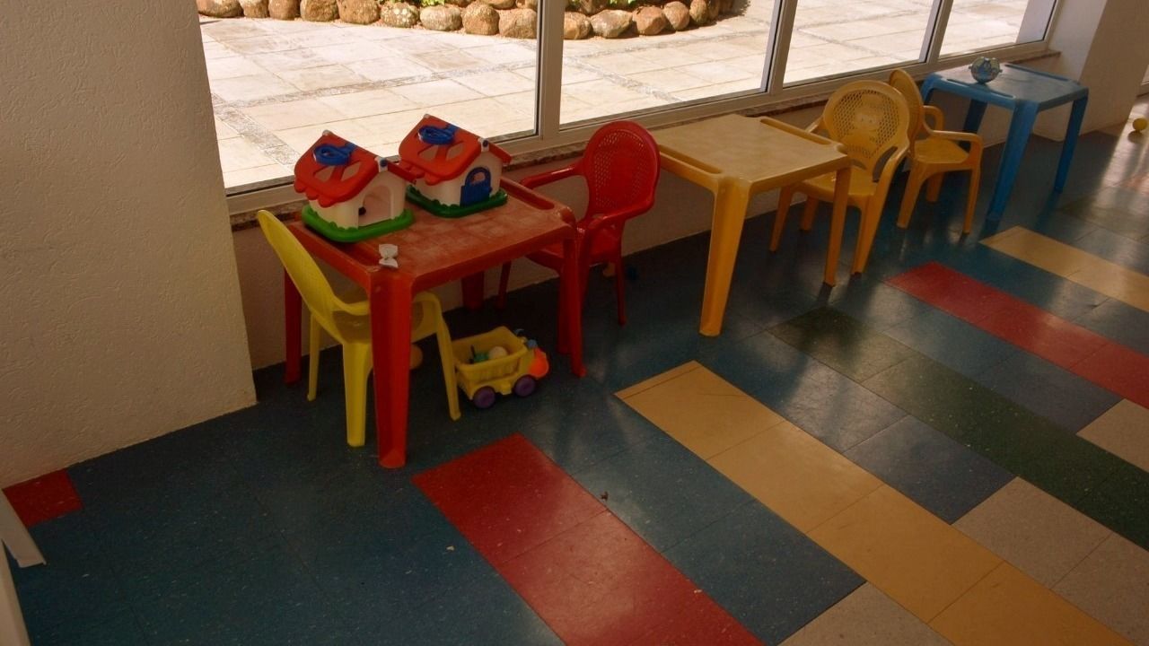 Espaço de recreação infantil com tapetes coloridos, uma mesa, cadeiras e brinquedos.