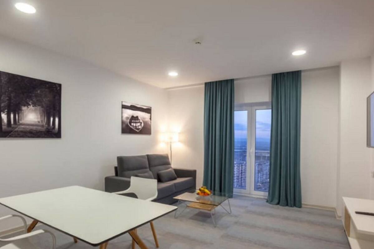 Een moderne woonkamer met een bank, een eettafel en een salontafel. Twee kunstwerken hangen aan de muur, een raam met gordijnen en uitzicht op de stad.