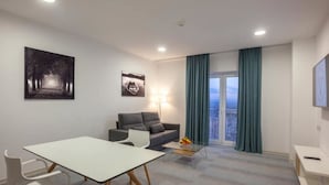 Un salon moderne avec un canapé, une table à manger et une table basse. Deux œuvres d’art ornent le mur, il y a une fenêtre avec des rideaux et une vue sur la ville.