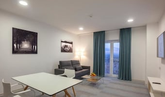 Una sala de estar moderna con un sofá, una mesa de comedor y una mesa de centro. Hay dos obras de arte en la pared, una ventana con cortinas y vistas a la ciudad.