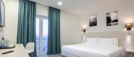 A hotel room with a bed, a desk, a chair, a window with curtains, and wall-mounted lamps.