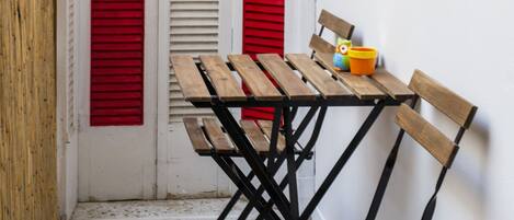 Een klein terras met een houten tafel en bankjes, omgeven door witte muren met rode luiken.