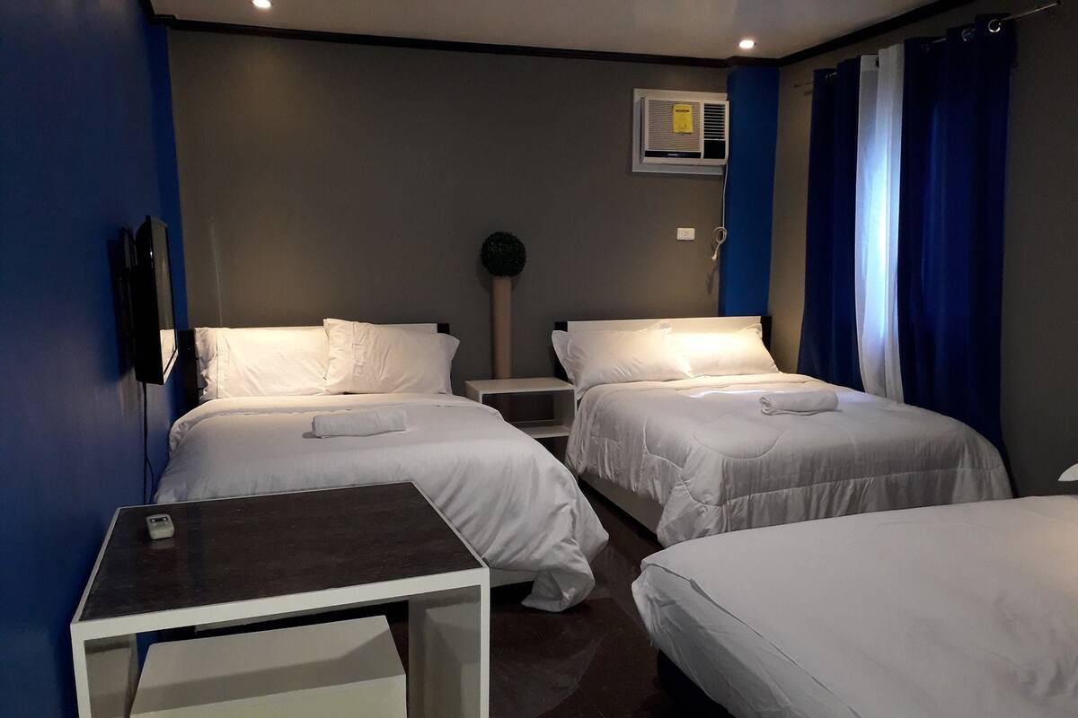 A hotel room with two single beds, a small table, a wall-mounted air conditioner, and a TV.