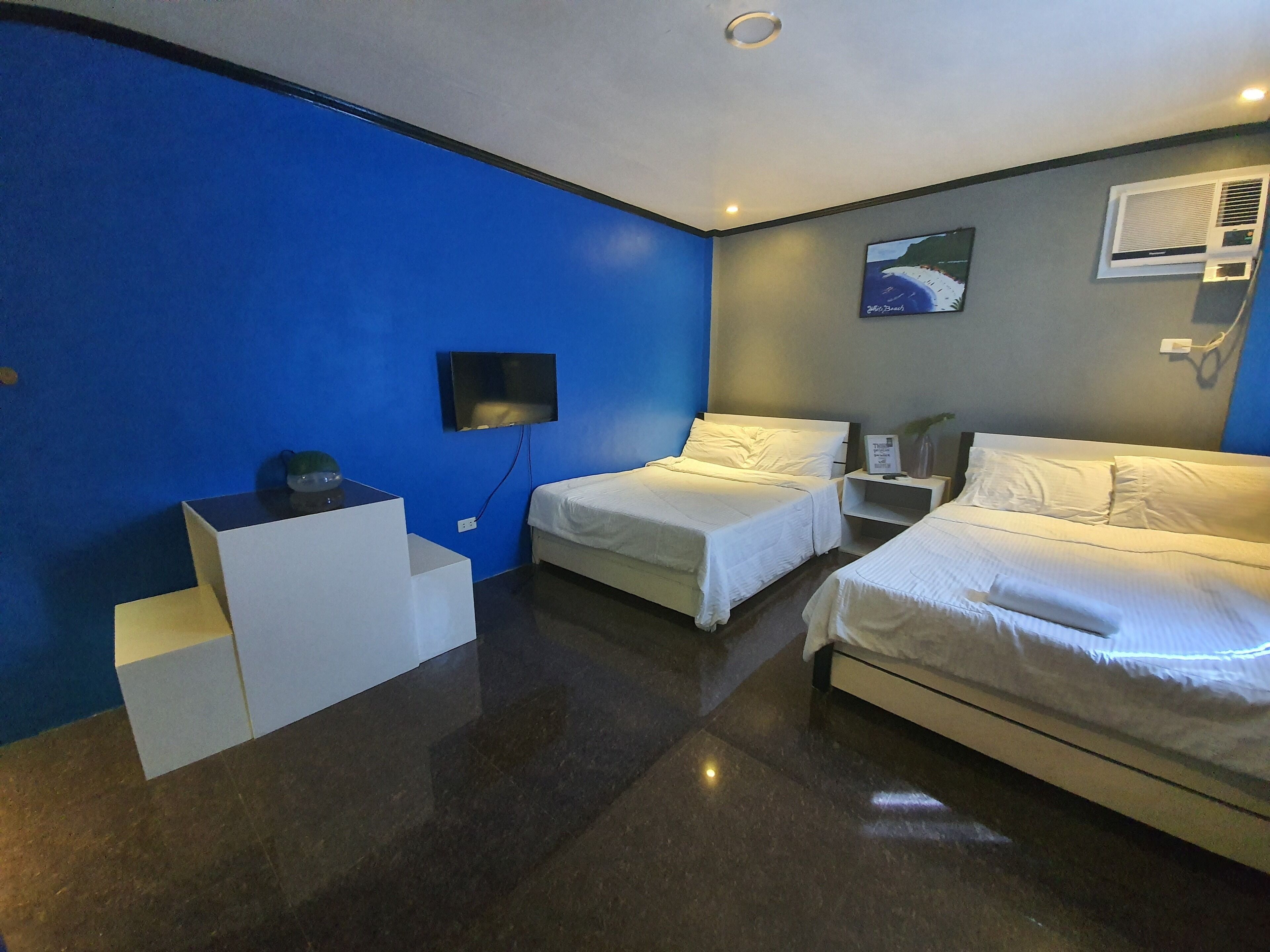 Habitación de hotel con dos camas, un televisor de pantalla plana montado en la pared, una mesita blanca y una pared decorada en azul.