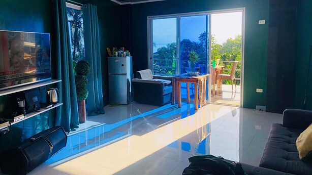 A modern living room with a flat-screen TV, a sofa, a coffee table, and a balcony with a view of greenery.