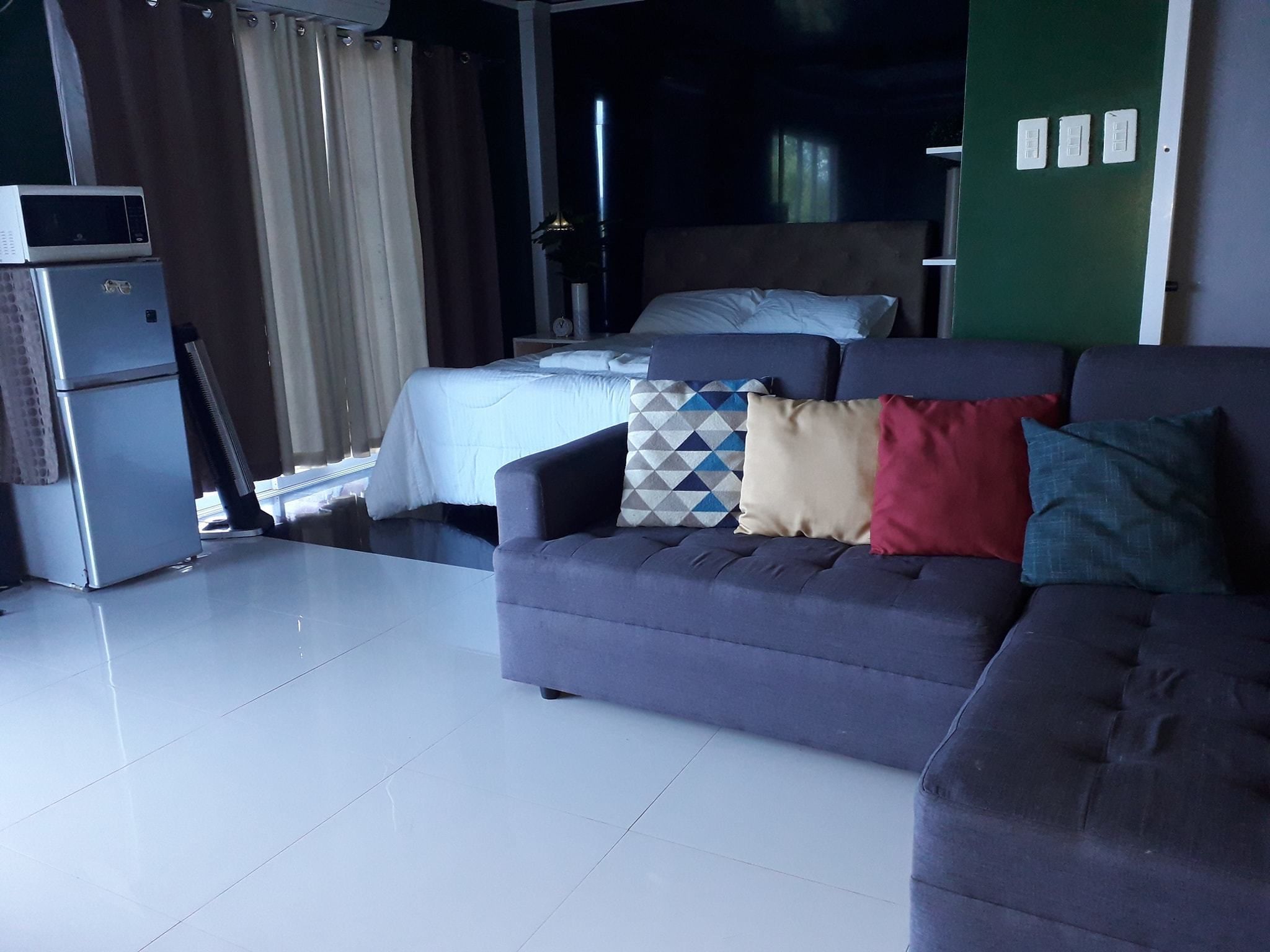 Una habitación de hotel moderna con un sofá gris, una cama con ropa blanca, una kitchenette pequeña y una pared de acento verde.