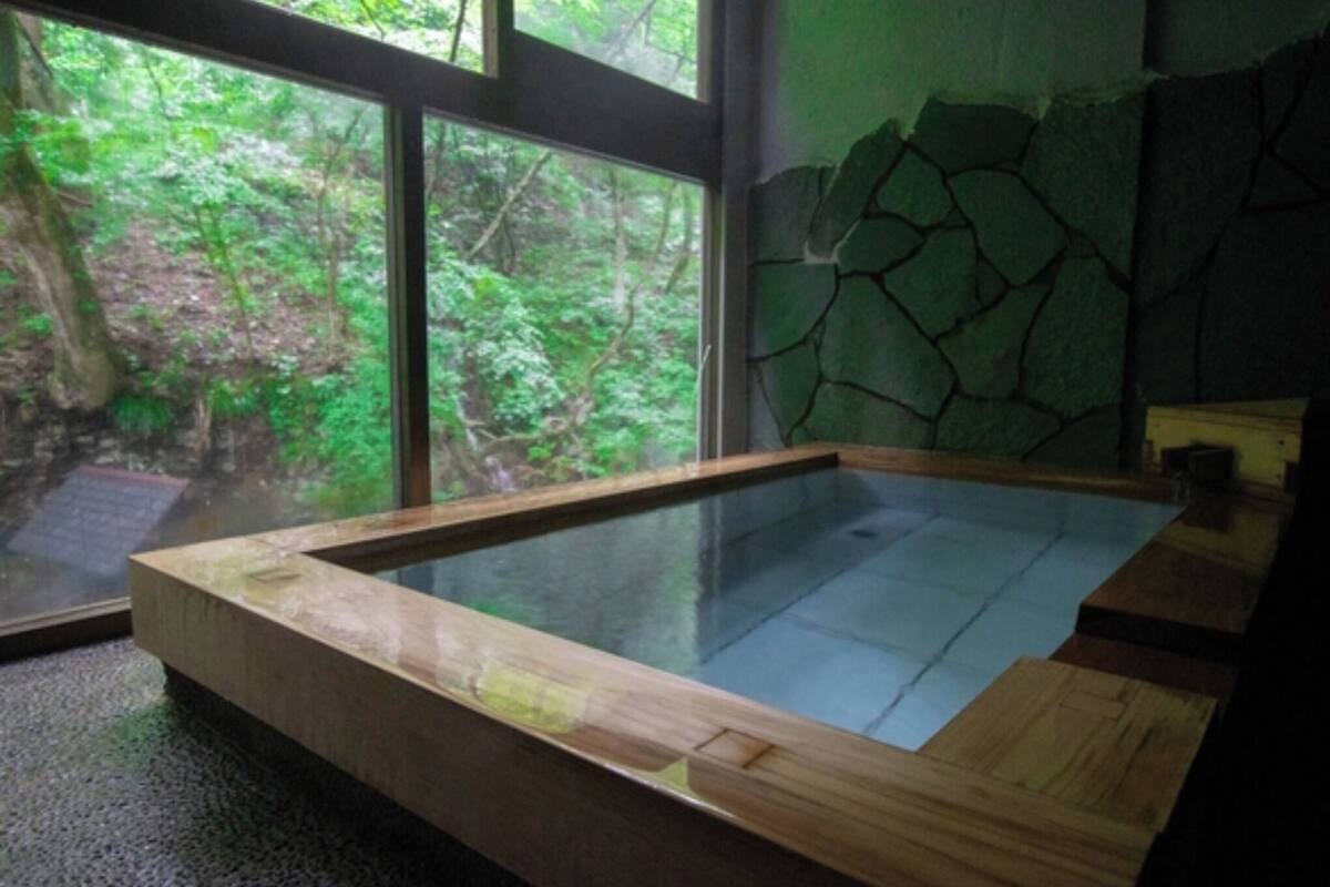 A wooden hot tub with a glass bottom, set against a stone wall and large windows overlooking a forest.