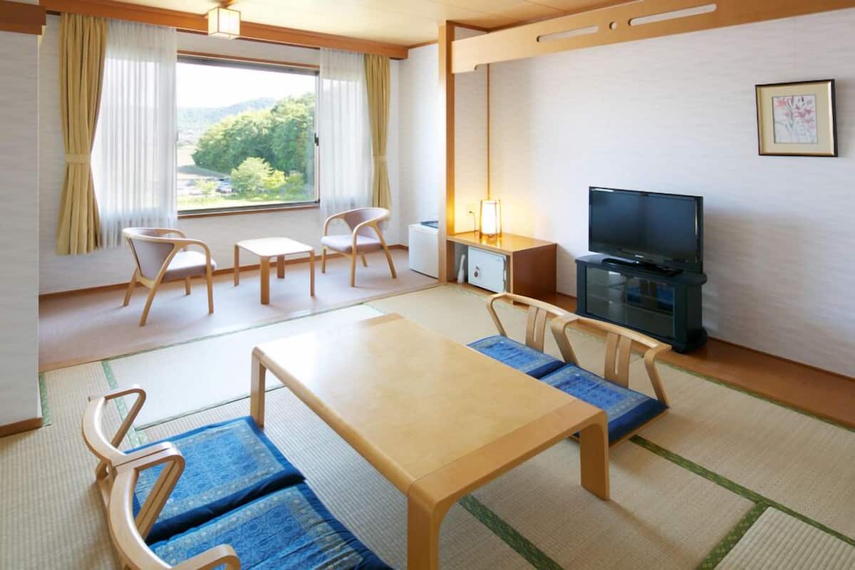 Ein Zimmer mit Tatami-Matten, einem Holztisch, Stühlen, einem Fernseher und einem Fenster mit Vorhängen.