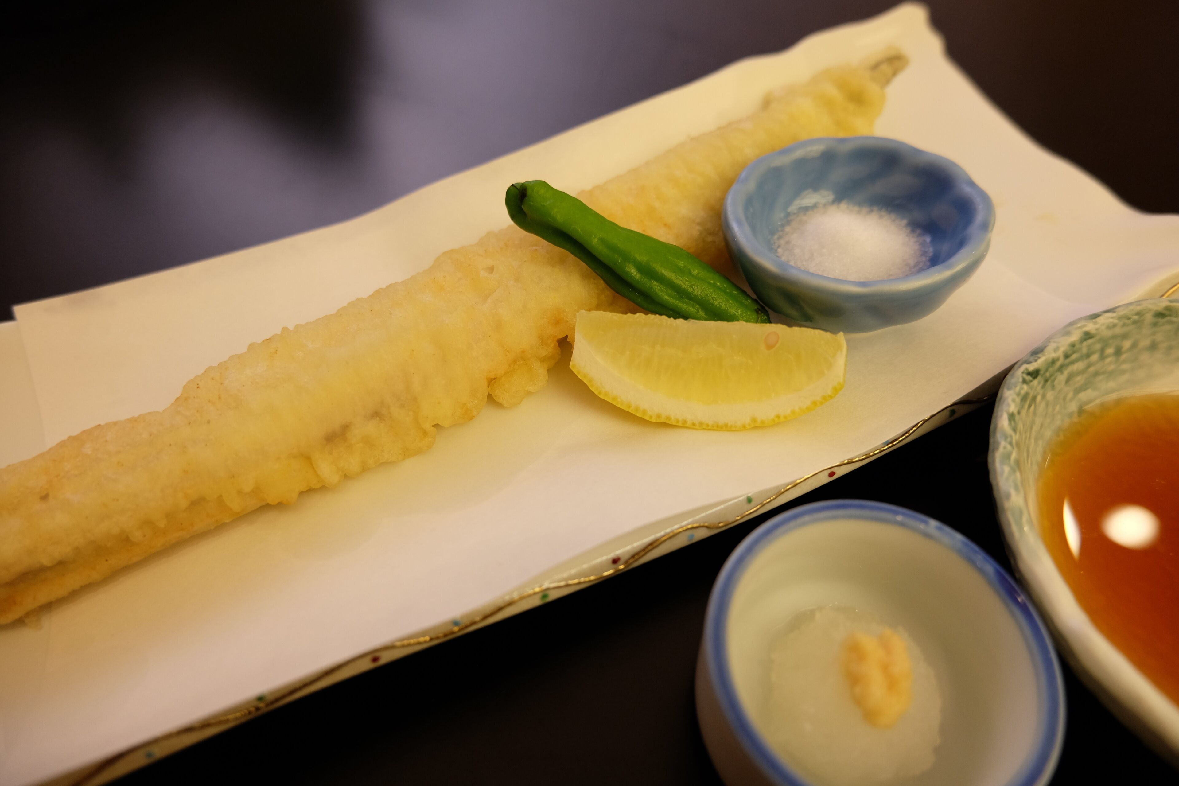 A plate with a single piece of fried food, a lemon wedge, and two small bowls of condiments.