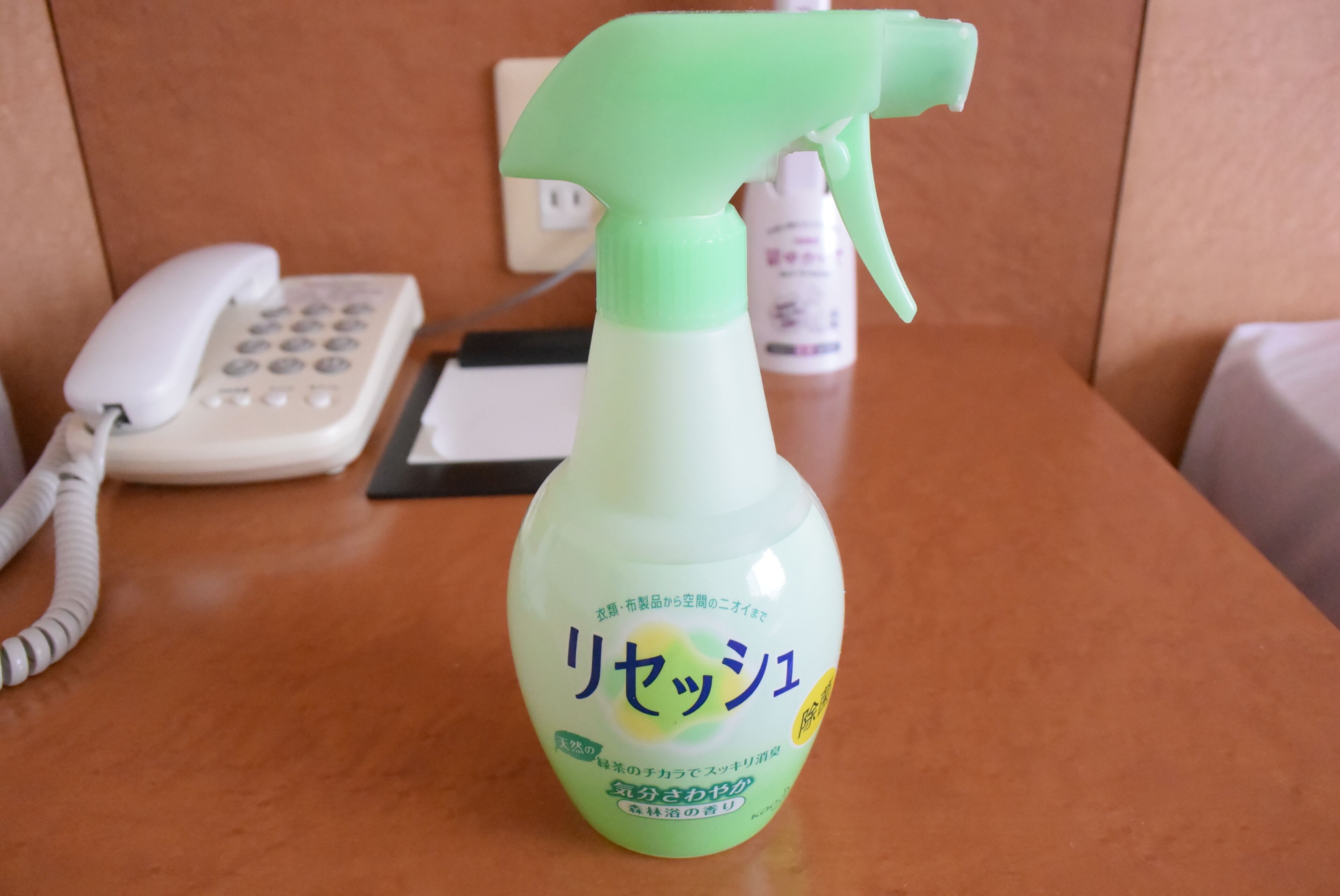 A spray bottle with Japanese text on a wooden surface, a white telephone, and a black object in the background.