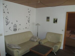 A living room with a beige sofa, a matching armchair, and a small wooden coffee table. There are two framed pictures on the wall and a lamp in the corner. - Ferienwohnungen Wiesenhof (Braunlage)