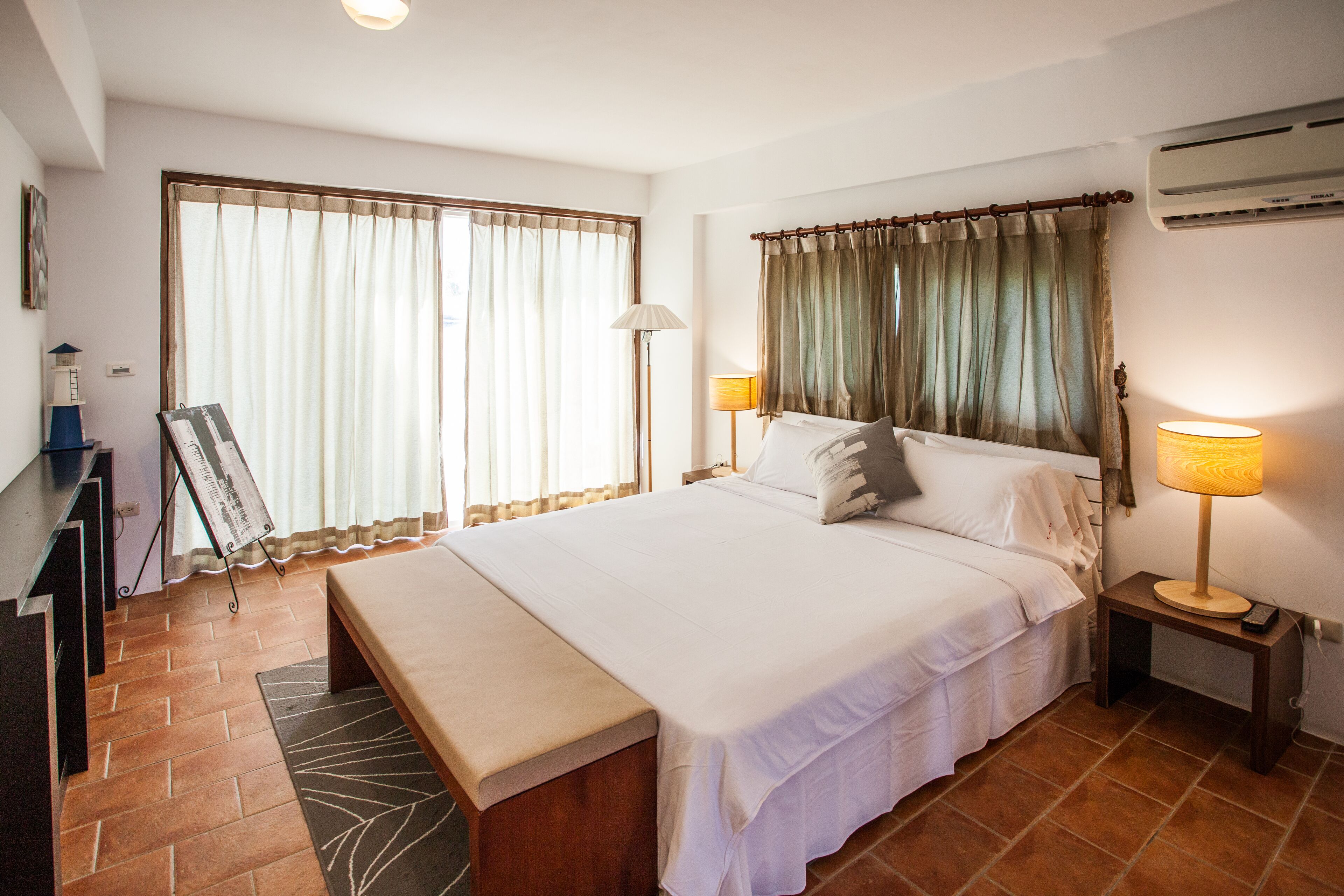 A hotel room with a large bed, a desk with a chair, a bench, a standing lamp, and a large window with curtains.