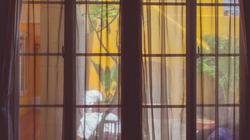 Una ventana con cortinas translúcidas, vista a una pared amarilla y una planta en maceta afuera.