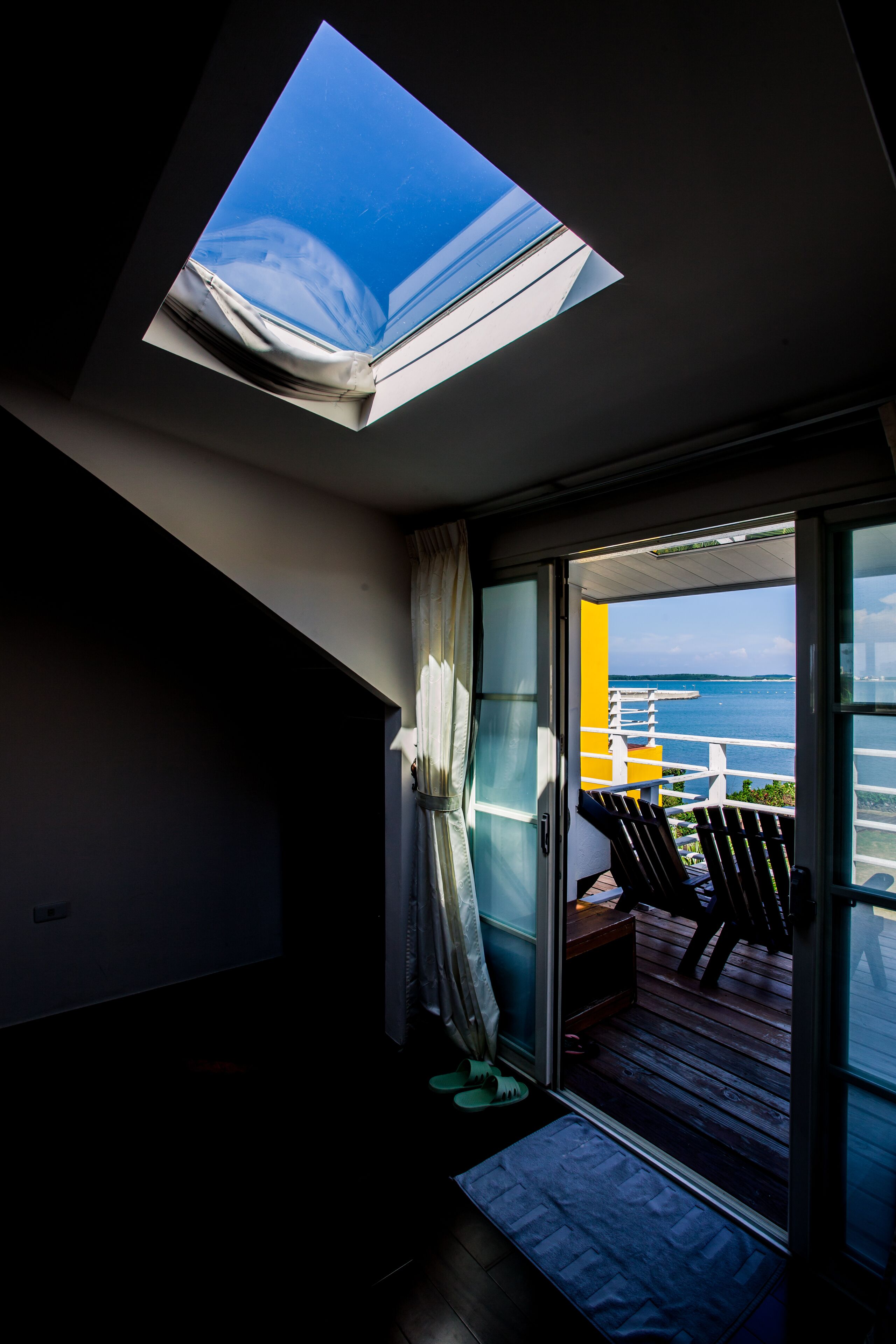 A room with a skylight, a sliding glass door leading to an outdoor deck with chairs, and a view of the sea.