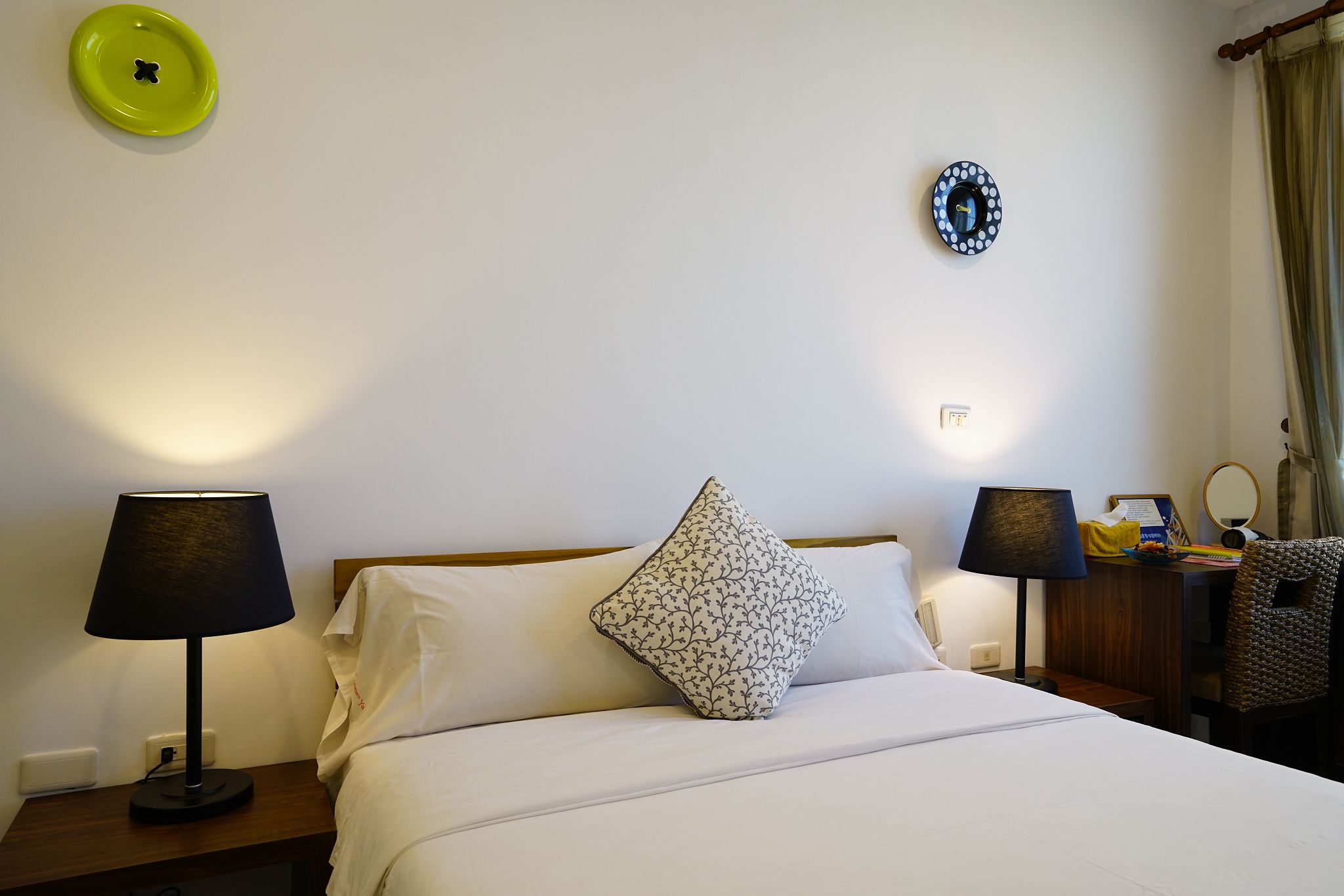 Una habitación de hotel con una cama, dos lámparas de noche, una mesita de noche, un reloj de pared y una pequeña mesa con un espejo y una caja.