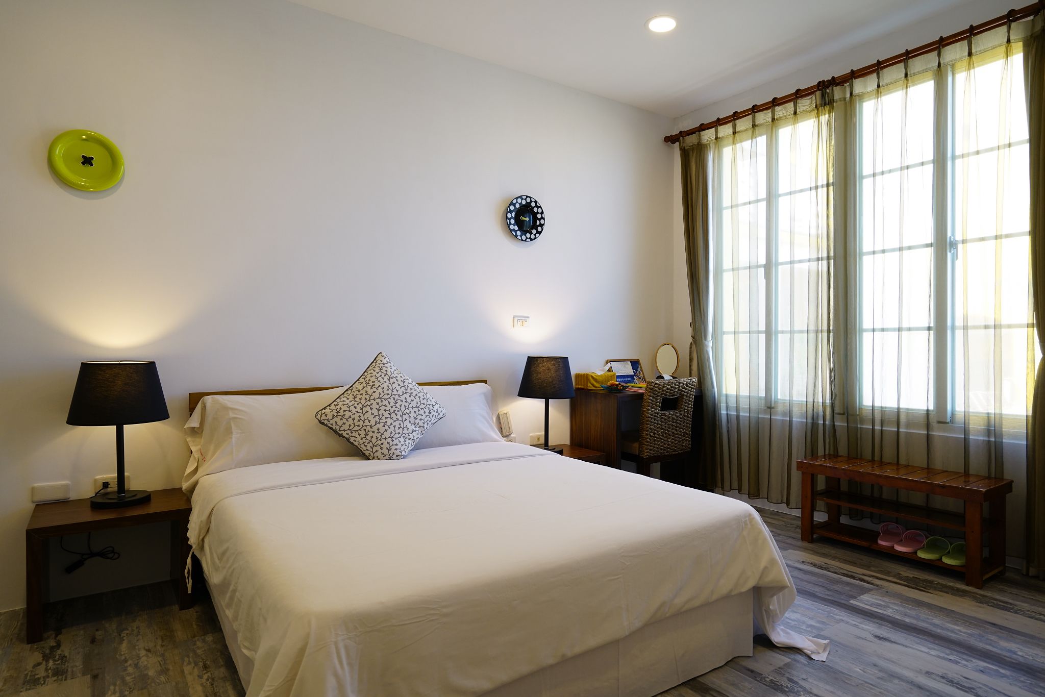 A neatly arranged bedroom with a bed, two bedside tables with lamps, a bedside clock, a bench with shoes, and a window with curtains.