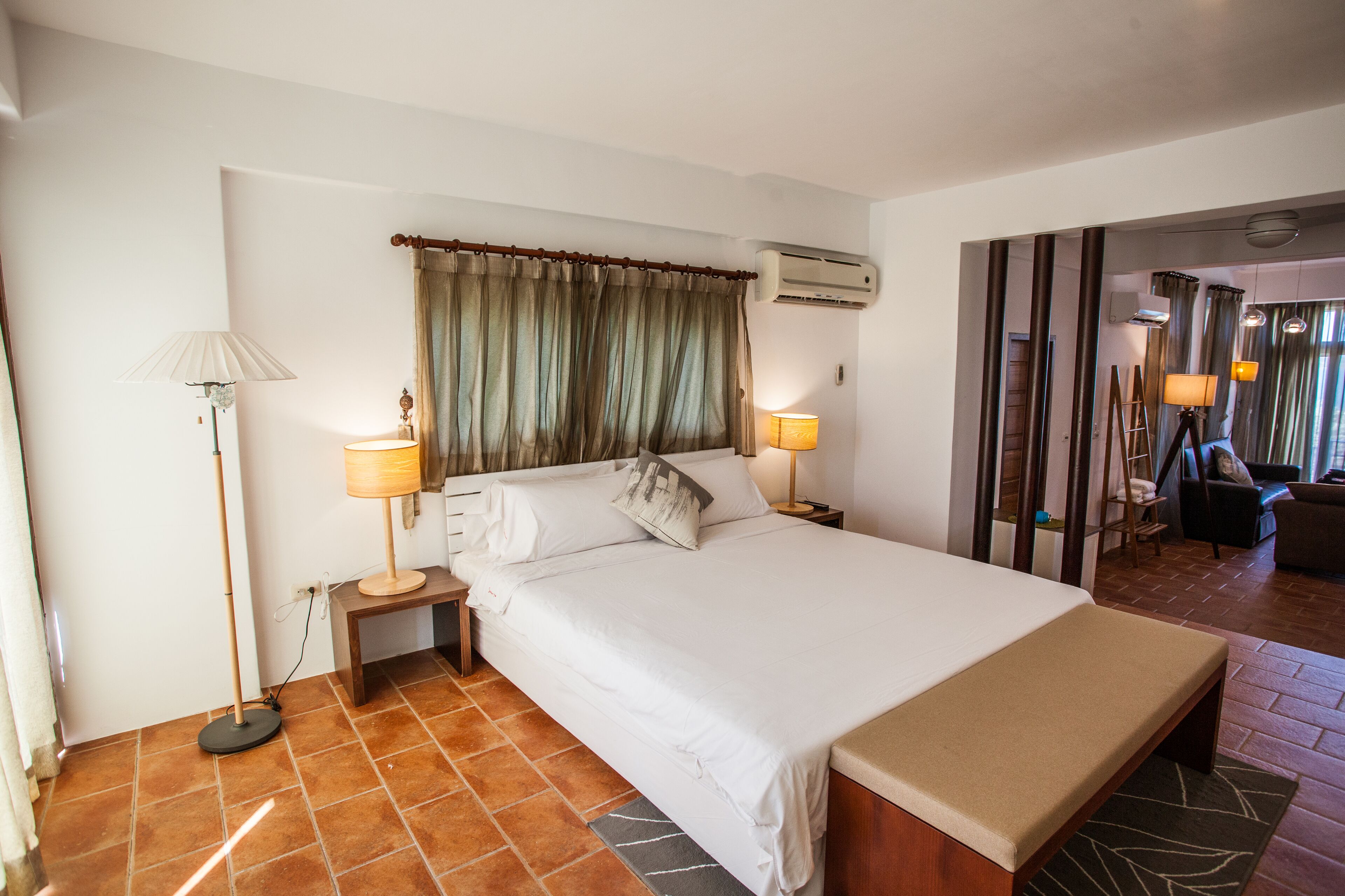 A hotel room with a large bed, bedside lamps, an air conditioning unit, and a view of the adjacent living area.