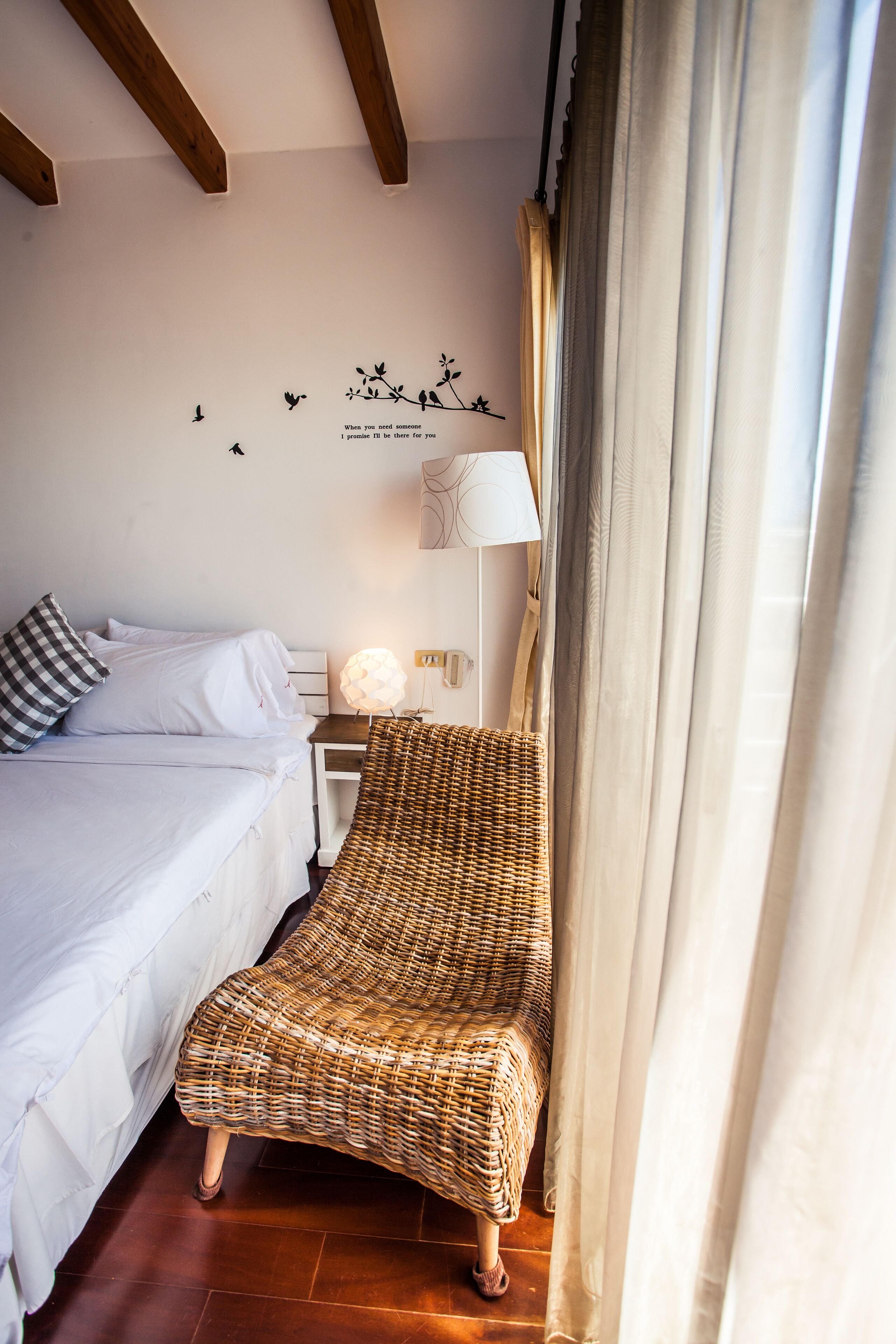 Een slaapkamer met een bed, een rieten stoel, een nachtkastje met een lamp en een muursticker.