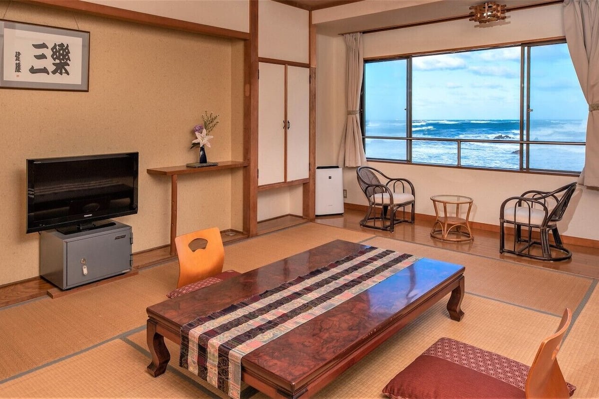 A room with a TV, a wooden table, chairs, a view of the ocean, and a tatami mat floor.