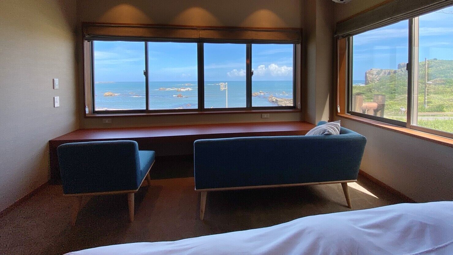 Une pièce avec un lit, deux chaises bleues, une fenêtre donnant sur la mer et un banc en bois.