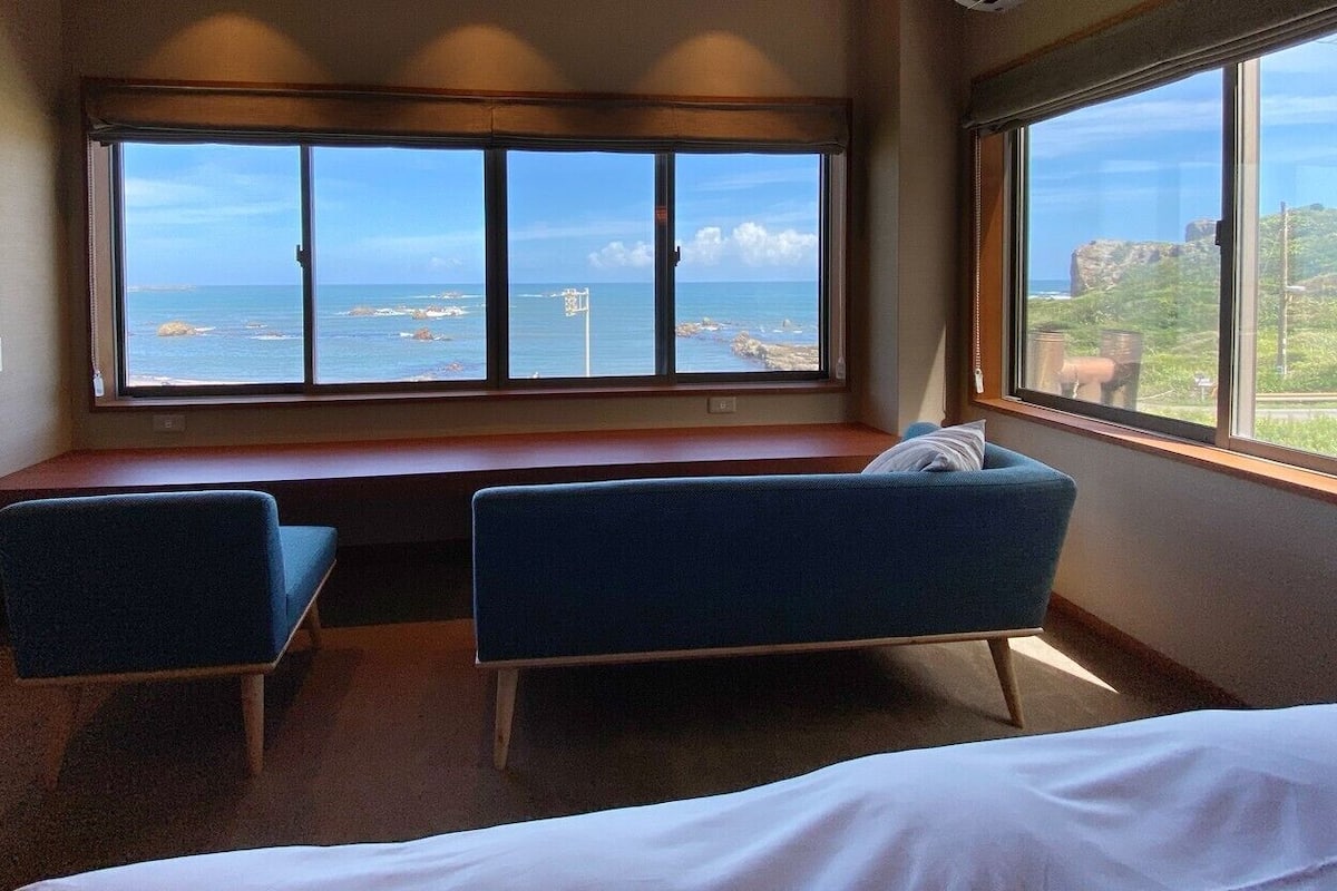 A room with a bed, two blue chairs, a window with a view of the sea, and a wooden bench.