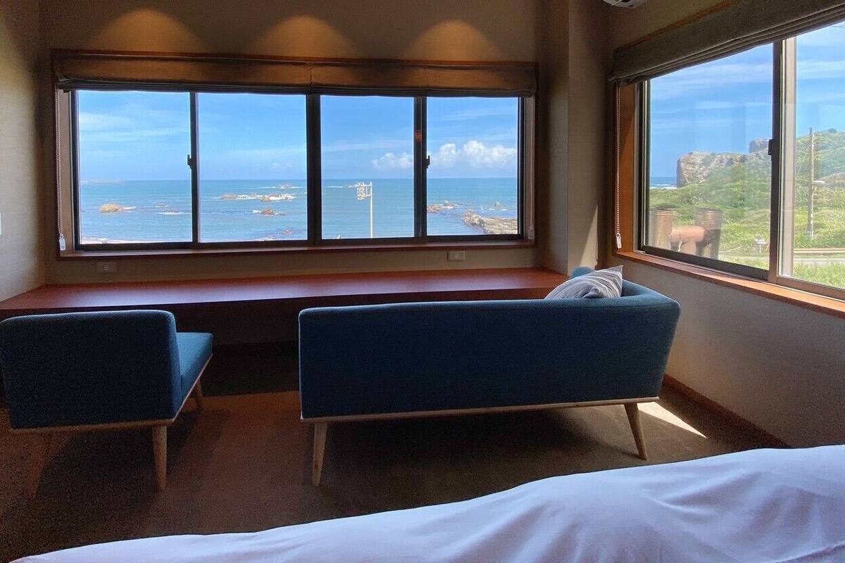 Une pièce avec un lit, deux chaises bleues, une fenêtre donnant sur la mer et un banc en bois.