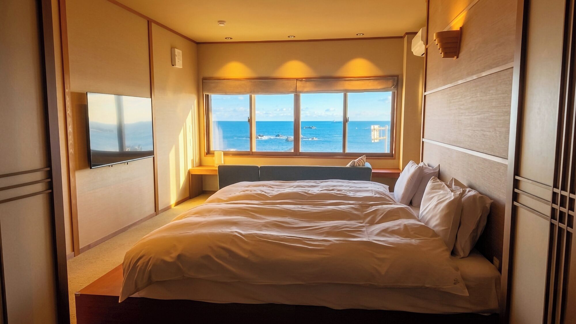 Habitació d'hotel amb llit, escriptori, cadira, finestra i vistes al mar, i televisió penjada a la paret.