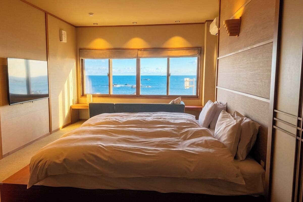 A hotel room with a bed, a desk, a chair, a window with a view of the sea, and a TV on the wall.