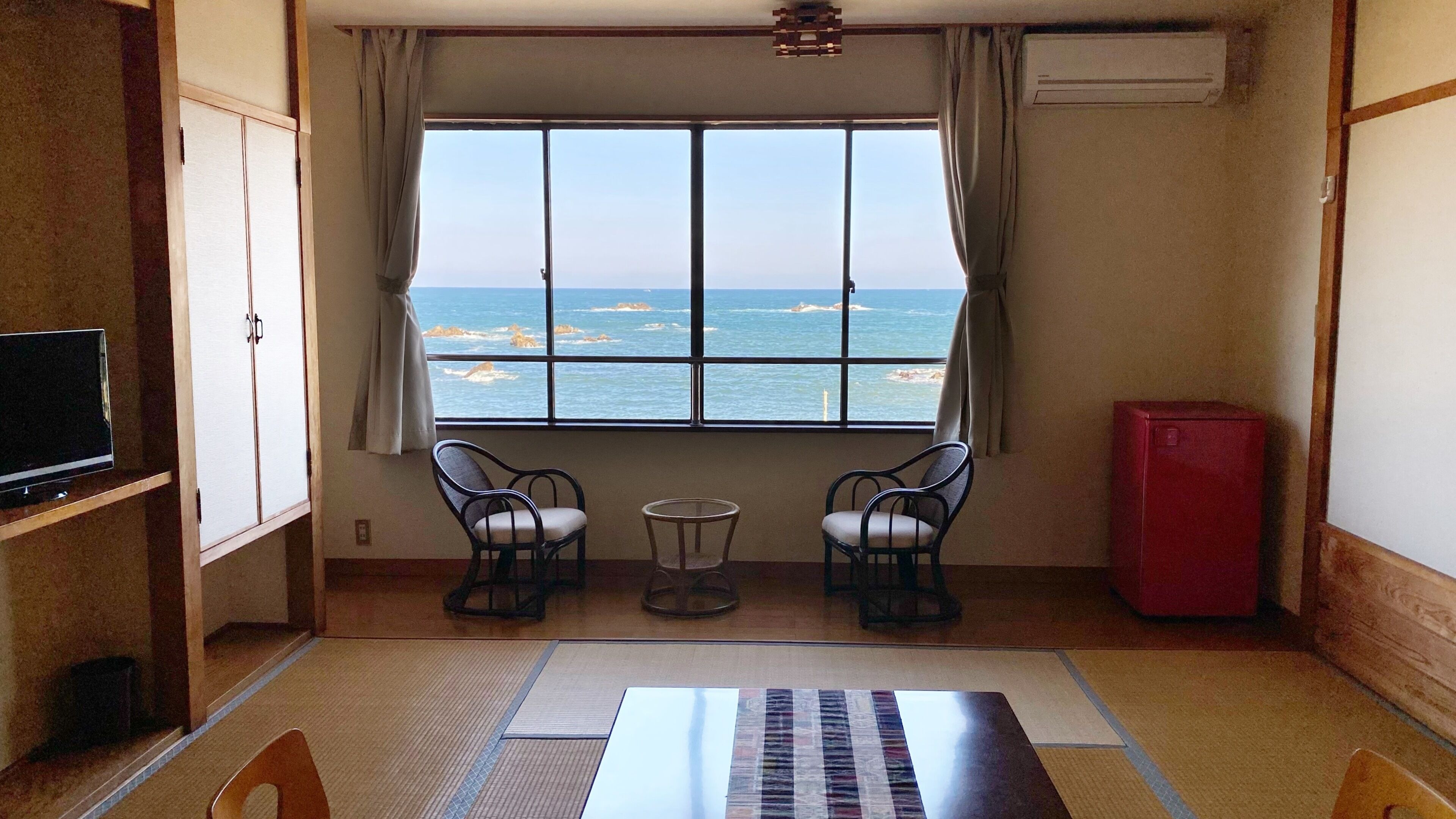 Une chambre avec vue sur la mer, deux fauteuils, une petite table, un téléviseur et un buffet rouge.
