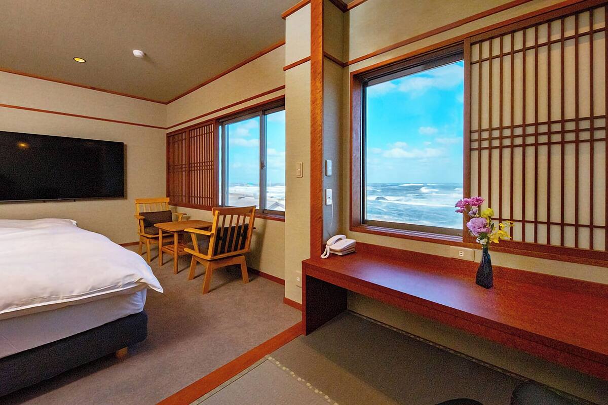 A hotel room with a bed, a desk, a chair, a phone, a vase with flowers, and a view of the ocean through a window with wooden slats.