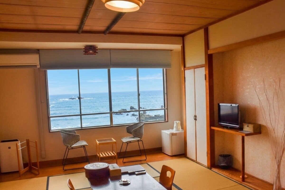 A room with a large window overlooking the ocean, a television on a shelf, and a traditional Japanese-style table with chairs.