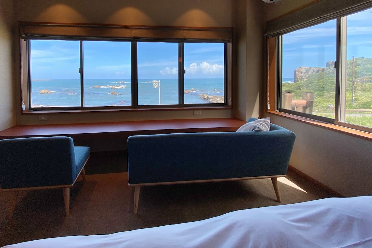 A hotel room with a large window, two blue chairs, a bed, and a wooden desk.