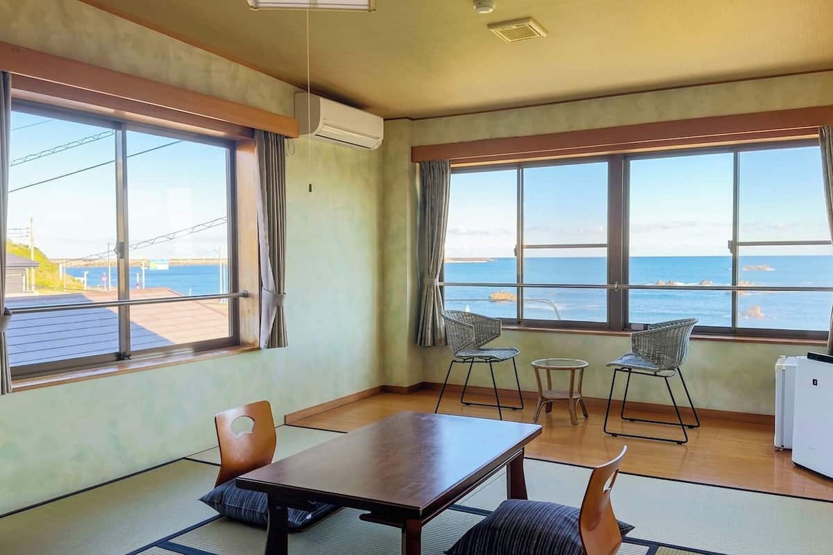 A room with a wooden table, two chairs, a small table, and a refrigerator. Large windows offer a view of the sea and a distant shoreline.
