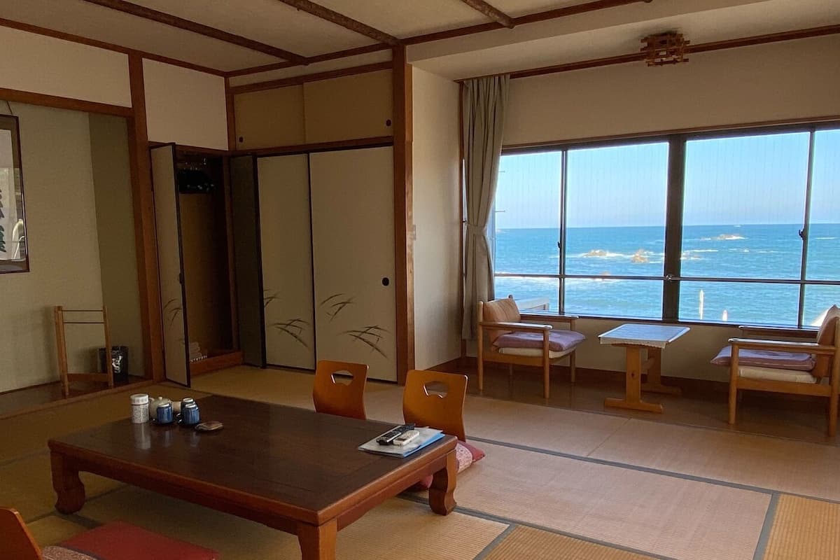 A traditional Japanese room with tatami flooring, a low table, and chairs. A large window offers a view of the ocean.
