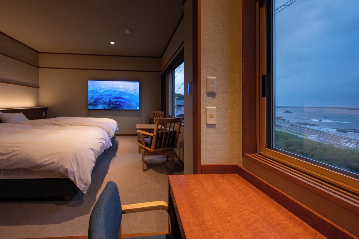 A hotel room with a bed, a desk, a chair, a TV, and a large window with a view of the sea.